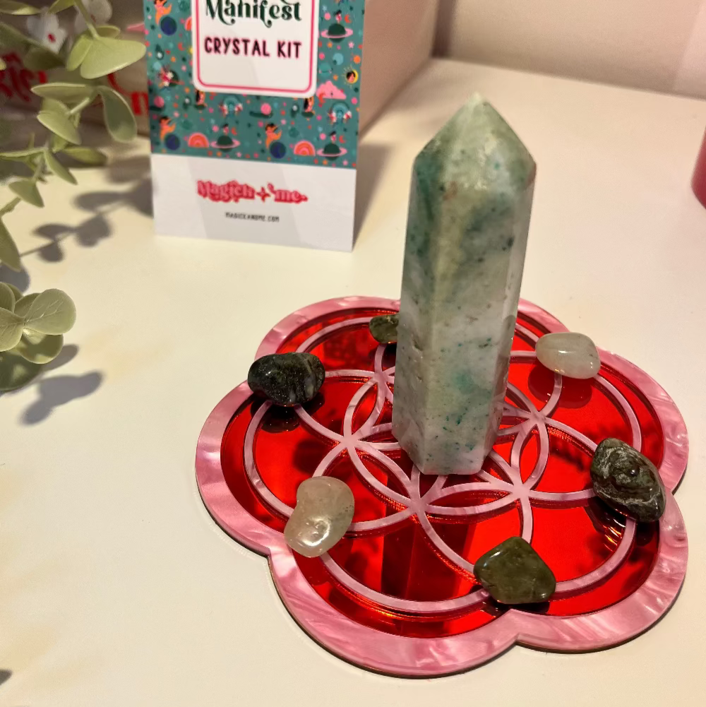 A red and pink crystal grid board with a Seed of Life design, featuring a central crystal and surrounded by smaller stones, displayed at a table with a colorful backdrop.
